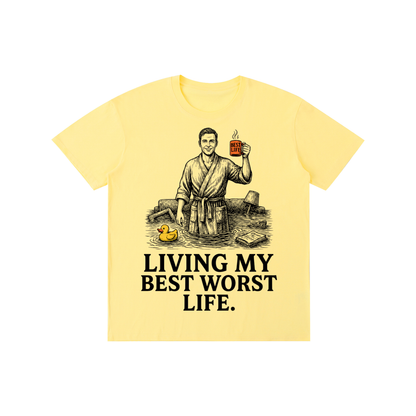 Yellow cotton crewneck t-shirt with a vintage illustration of a smiling man in a bathrobe raising a mug in a flooded room beside a rubber duck above bold Living my best worst life text on the front