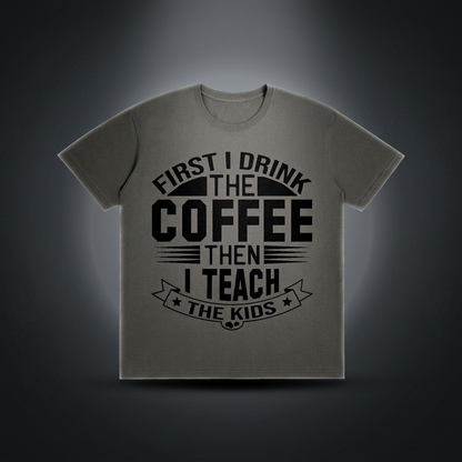 Dark gray cotton crewneck t-shirt with black First I drink the coffee then I teach the kids lettering with a star ribbon banner on the front shown against a dark studio backdrop