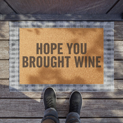 Hope You Brought Wine coir doormat on a wooden porch seen from above with a pair of shoes at the edge