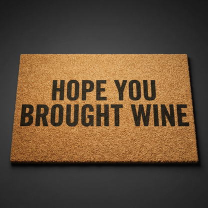 Hope You Brought Wine coir doormat with dark lettering on natural tan coir, shown at an angle on a dark studio backdrop
