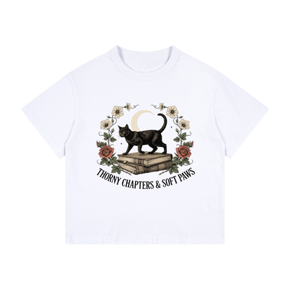 Kids’ loose-fit drop-shoulder tee with a black cat walking over old books under a crescent moon, Thorny Chapters & Soft Paws front print