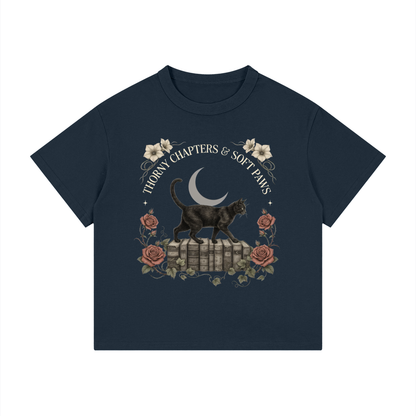 Kids’ loose-fit drop-shoulder tee with a black cat walking over old books under a crescent moon, Thorny Chapters & Soft Paws front print