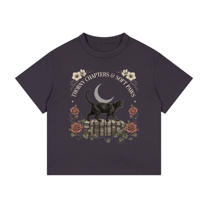 Kids’ loose-fit drop-shoulder tee with a black cat walking over old books under a crescent moon, Thorny Chapters & Soft Paws front print