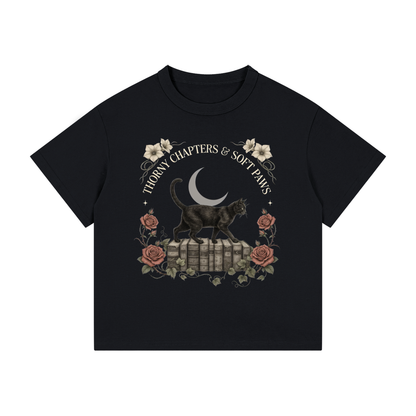 Kids’ loose-fit drop-shoulder tee with a black cat walking over old books under a crescent moon, Thorny Chapters & Soft Paws front print