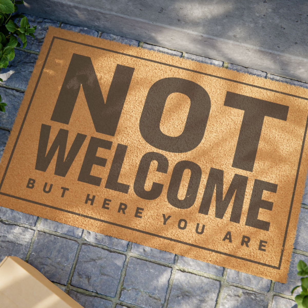 Close view of the Not Welcome But Here You Are coir doormat on a stone patio in dappled sunlight