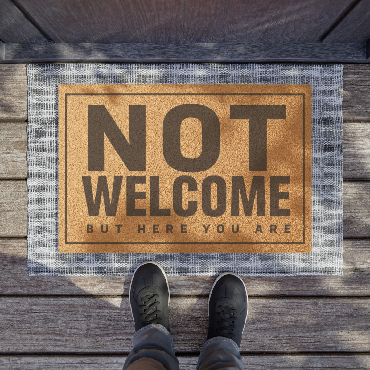 Not Welcome But Here You Are coir doormat on a wooden porch seen from above with a pair of shoes at the edge