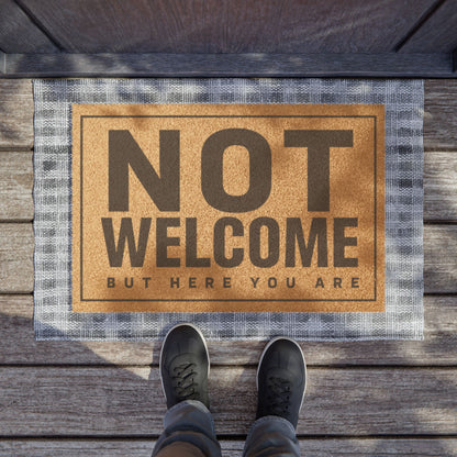 Not Welcome But Here You Are coir doormat on a wooden porch seen from above with a pair of shoes at the edge