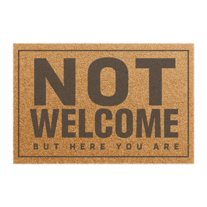 Top view of the Not Welcome But Here You Are coir doormat, dark lettering inside a thin border on natural tan fiber