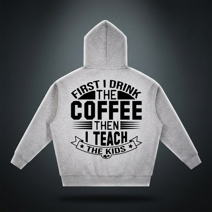 Heather gray oversized boxy fleece hoodie with black First I drink the coffee then I teach the kids lettering with a star ribbon banner on the back shown against a dark studio backdrop