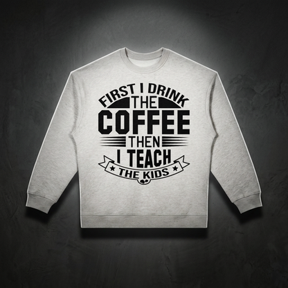 Heather white oversized crewneck sweatshirt with black First I drink the coffee then I teach the kids lettering with a star ribbon banner on the front shown against a dark studio backdrop