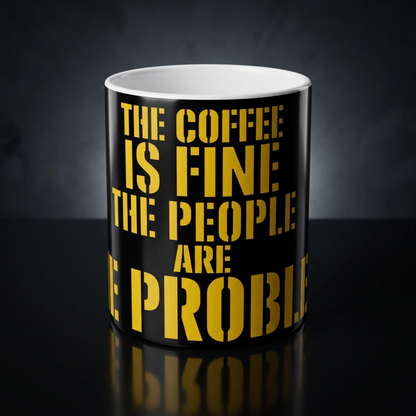 Funny coffee mug printed with "THE COFFEE IS FINE THE PEOPLE ARE THE PROBLEM", shown on a dark studio background