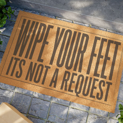 Close view of the Wipe Your Feet It's Not a Request coir doormat on a stone patio in dappled sunlight