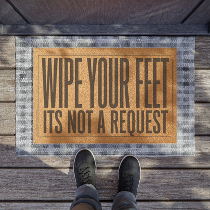 Wipe Your Feet It's Not a Request coir doormat on a wooden porch seen from above with a pair of shoes at the edge