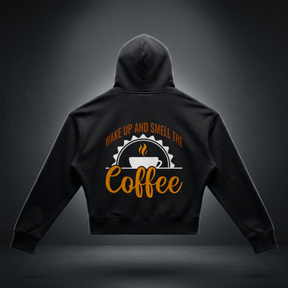 Black women's cropped drop shoulder hoodie with a rust brown Wake up and smell the coffee design with a white coffee cup and orange cursive Coffee on the back shown against a dark studio backdrop