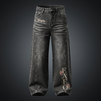 Black vintage wash wide leg jeans with FAFO lettering and pink flowers on the lower leg, shown against a dark studio backdrop