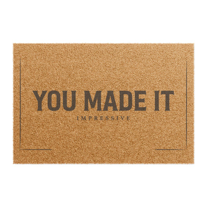 Top view of the You Made It Impressive coir doormat, dark lettering inside a thin border on natural tan fiber
