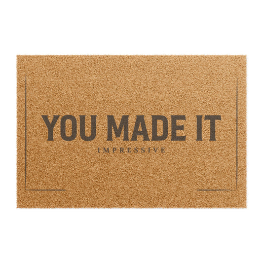 Top view of the You Made It Impressive coir doormat, dark lettering inside a thin border on natural tan fiber