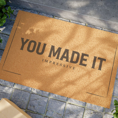 Close view of the You Made It Impressive coir doormat on a stone patio in dappled sunlight